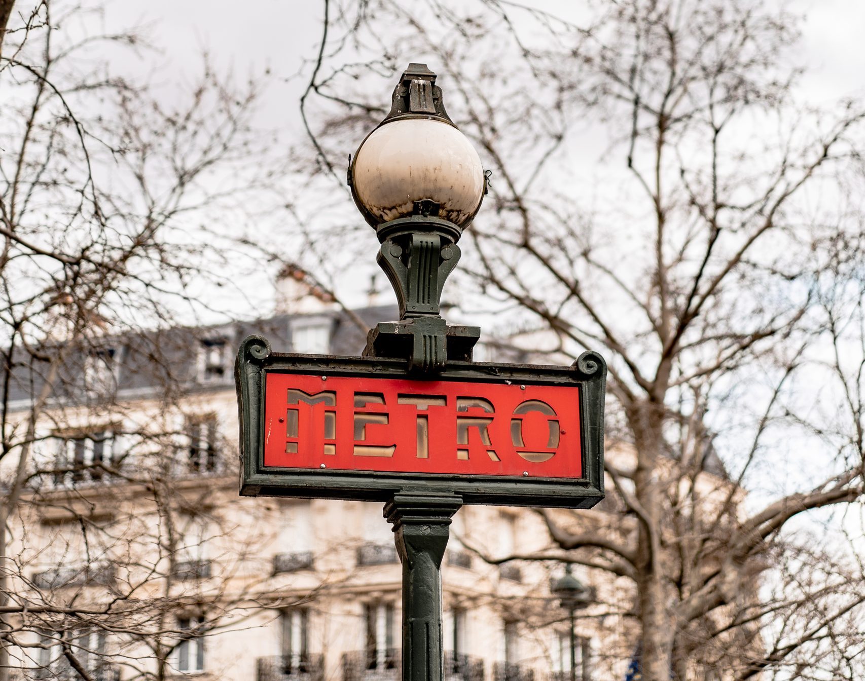 Parīzes metro zīme ar sarkanu uzrakstu “Métro” uz laternas, fonā kaili koki