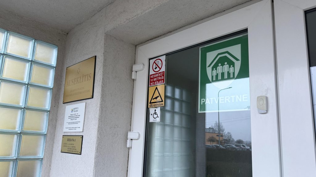 Tuvplāns ēkas ieejai ar informatīvām plāksnēm un zaļu norādi "PATVERTNE" uz stikla durvīm.