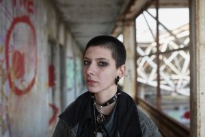 Jauna sieviete ar īsiem matiem un alternatīva stila apģērbu pozē industriālā ēkā ar grafiti uz sienām.