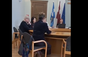 Grupa cilvēku sēž pie apaļa koka galda sapulču telpā; fonā redzami Rēzeknes pilsētas, Latvijas un Eiropas Savienības karogi.