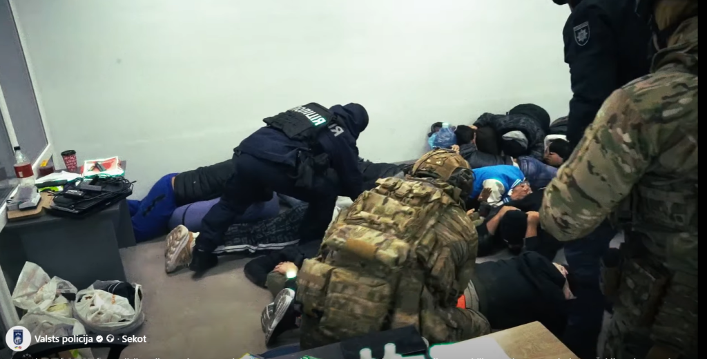 Bruņoti policisti specapģērbā uzrauga aizturētās personas zvanu centra telpās.