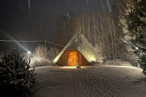 Svētes svētnīca ziemas naktī sniegotā ainavā ar izgaismotām durvīm.
