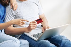Pāris sēž uz dīvāna un iepērkas internetā, izmantojot portatīvo datoru un bankas karti.