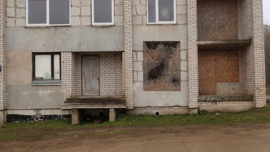 Tuvplāns uz veca daudzdzīvokļu nama pirmo stāvu, kur redzamas slēgtas durvis un aizmūrēts logs ar saplākšņa dēļiem.