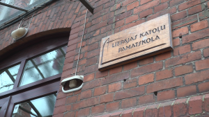 Brūna koka plāksne uz sarkanu ķieģeļu sienas ar uzrakstu "LIEPĀJAS KATOĻU PAMATSKOLA".