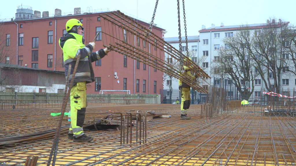 Strādnieks neona dzeltenā aizsargtērpā ar celtni paceļ lielu armatūras sietu Liepājas būvlaukumā.