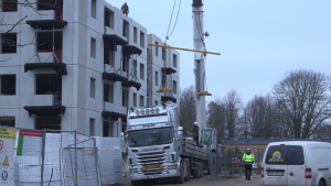 Celtnis paceļ liela izmēra dzelzsbetona sienas paneli uz jaunbūves augšējo stāvu Liepājā, blakus stāv kravas auto Scania.