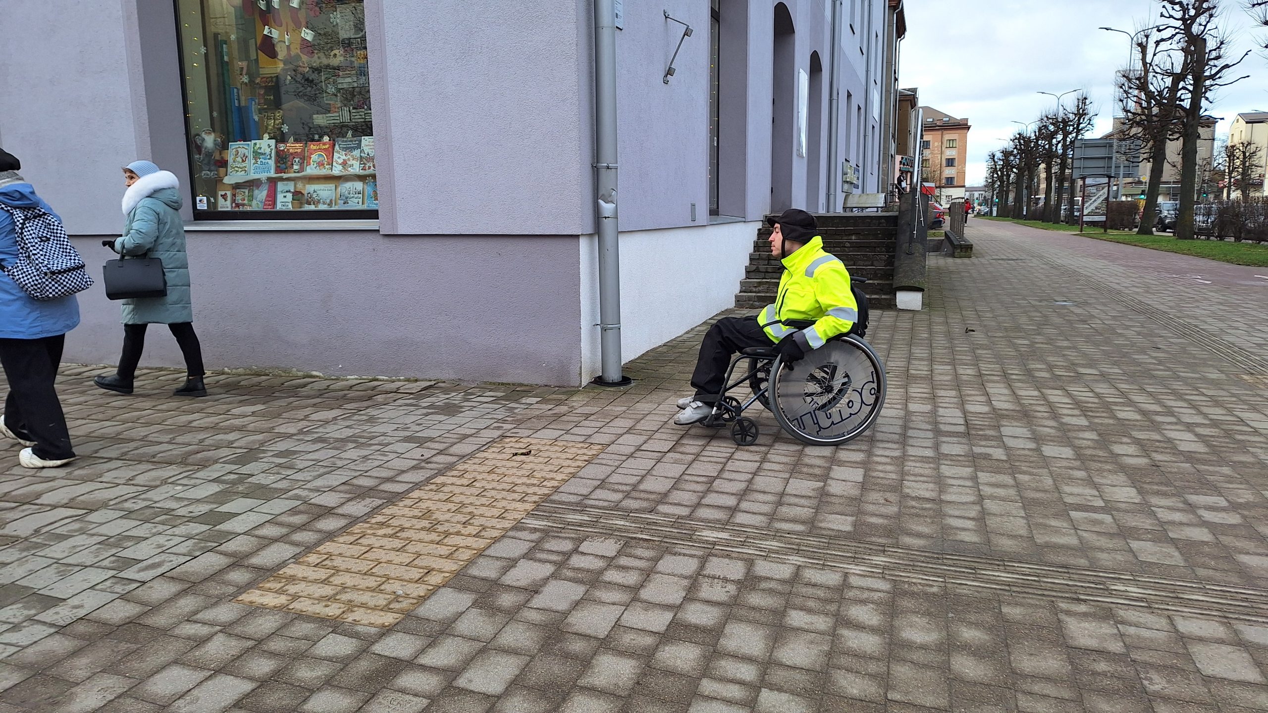 Cilvēks ratiņkrēslā uz ietves Jelgavā, izvērtējot pilsētvides pieejamību cilvēkiem ar kustību traucējumiem