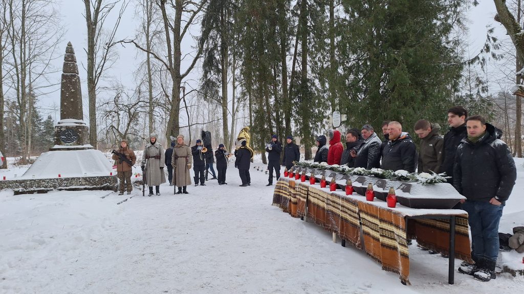 Cilvēku grupa un godasardze vēsturiskos tērpos stāv pie sniegota pieminekļa; priekšplānā uz galdiem rindā izvietoti mazi šķirsti ar svecēm.