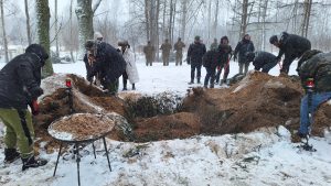 Cilvēki ziemas apģērbā ar lāpstām ber smiltis kopīgā kapu bedrē sniegotā mežā; priekšplānā svecītes un trauks ar smiltīm.