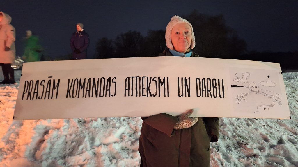 Sieviete ziemas apģērbā rokās tur garu, baltu plakātu ar tekstu "PRASĀM KOMANDAS ATTIEKSMI UN DARBU".