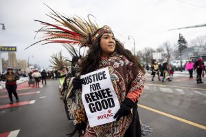 Sieviete tradicionālā apģērbā ar spalvu galvasrotu rokās tur plakātu "JUSTICE FOR RENEE GOOD".