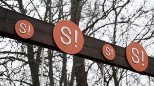 Koka konstrukcija parkā ar vairākiem oranžiem apļiem un uzrakstu “SI!”, fonā kaili koki ziemas sezonā
