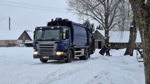 Atkritumu savākšanas automašīna sniegotā lauku teritorijā, darbinieks pie konteinera ziemas apstākļos.