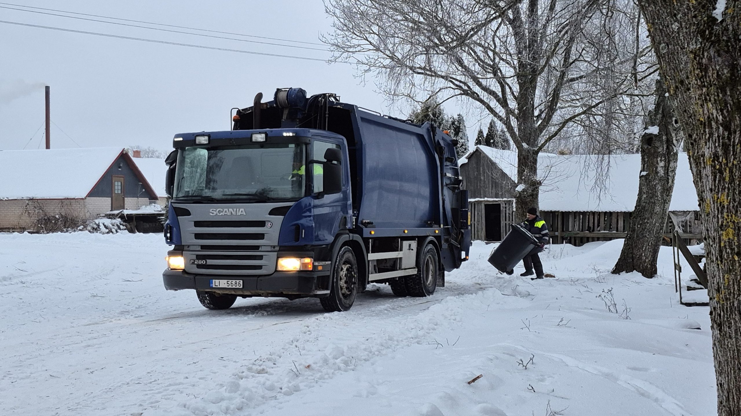Atkritumu savākšanas automašīna sniegotā lauku teritorijā, darbinieks pie konteinera ziemas apstākļos.