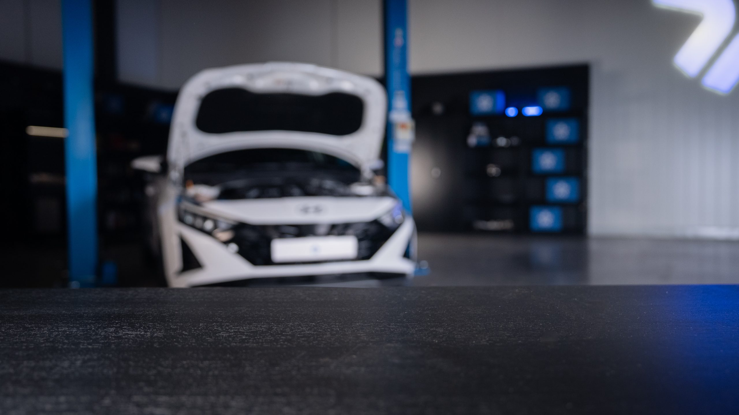 Automobiļa priekšpuse autoservisā uz pacēlāja, fonā redzama diagnostikas un riepu maiņas zona