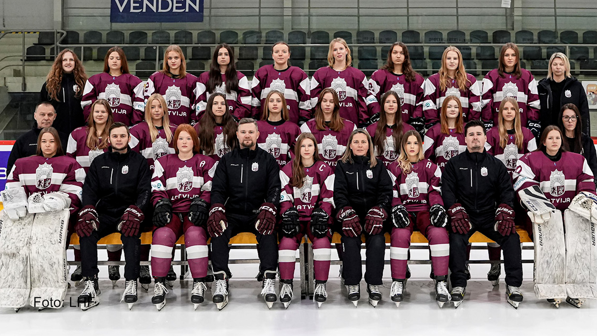 Latvijas U18 meiteņu hokeja izlase kopbildē uz ledus ar treneriem, gatavojoties pasaules čempionātam.