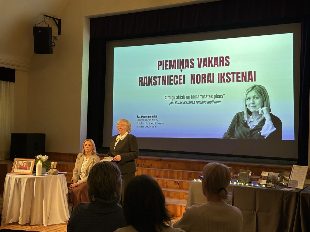 Dace Rempe uzstājas Noras Ikstenas piemiņas vakarā pie ekrāna ar rakstnieces portretu.
