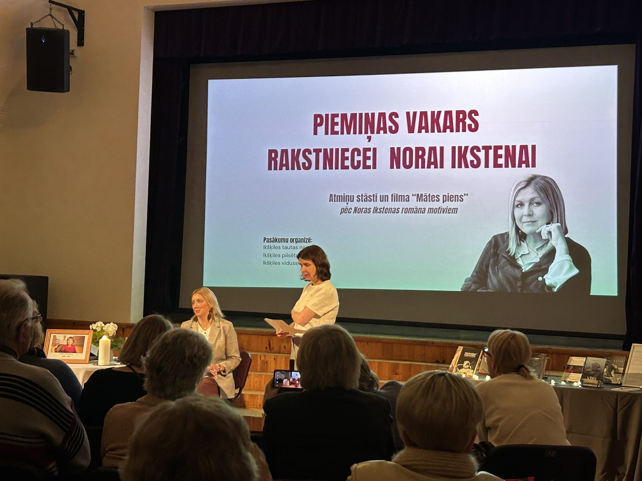 Piemiņas pasākums rakstniecei Norai Ikstenai Ikšķiles Tautas namā; uz skatuves ekrāna redzama rakstnieces fotogrāfija.