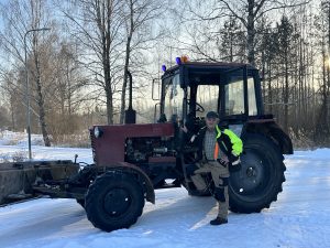 Komunālais darbinieks ziemā stāv pie traktora ar sniega lāpstu, veicot ceļu uzturēšanas darbus