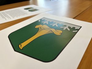 Uz galda novietots drukāts ģerboņa attēls ar stilizētu sēni un sniegpārslām uz zaļa fona