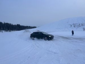 Tumši zaļa automašīna veic sānslīdi pa apsnigušu un aplejojošu trasi Jēkabpilī, aiz sevis paceļot sniega mākoni.