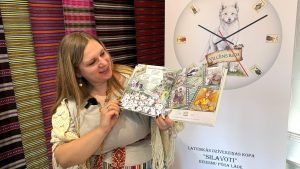 Sieviete tradicionālā tērpā prezentē ilustrētu bērnu grāmatu par latviešu mitoloģiju un gadskārtām