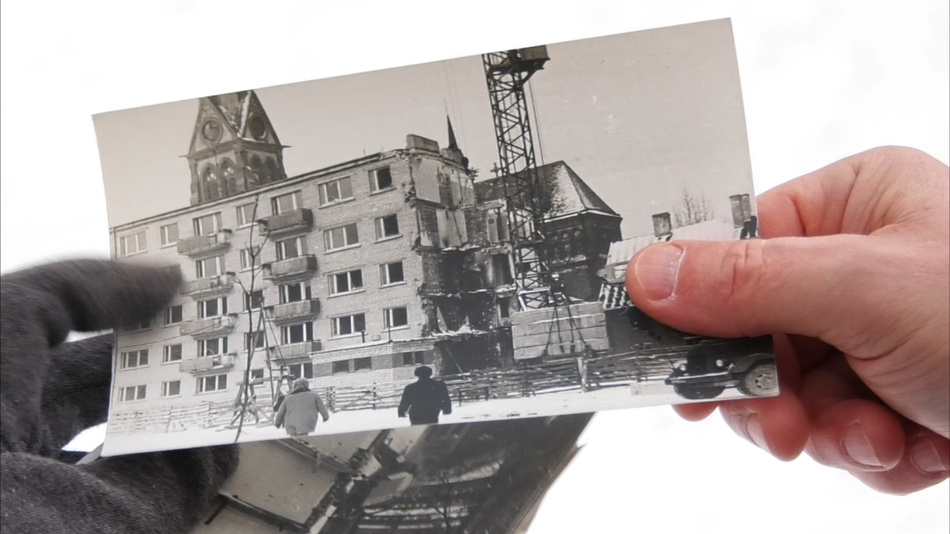 Cilvēka rokas tur melnbaltu fotogrāfiju, kurā redzama daļēji sagruvusi piecstāvu dzīvojamā māja Jelgavā pēc gāzes sprādziena.