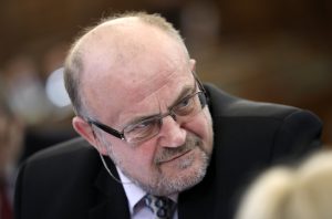 Bijušā Saeimas deputāta Jāņa Ādamsona portrets; vīrietis ar brillēm un uzvalku skatās sānis.