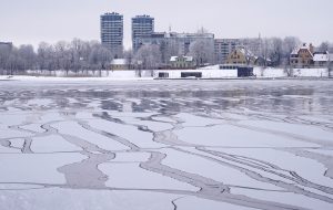Daugava Rīgā ziemā ar nevienmērīgu ledu un plaisām, fonā pilsētas ēkas un krasts.