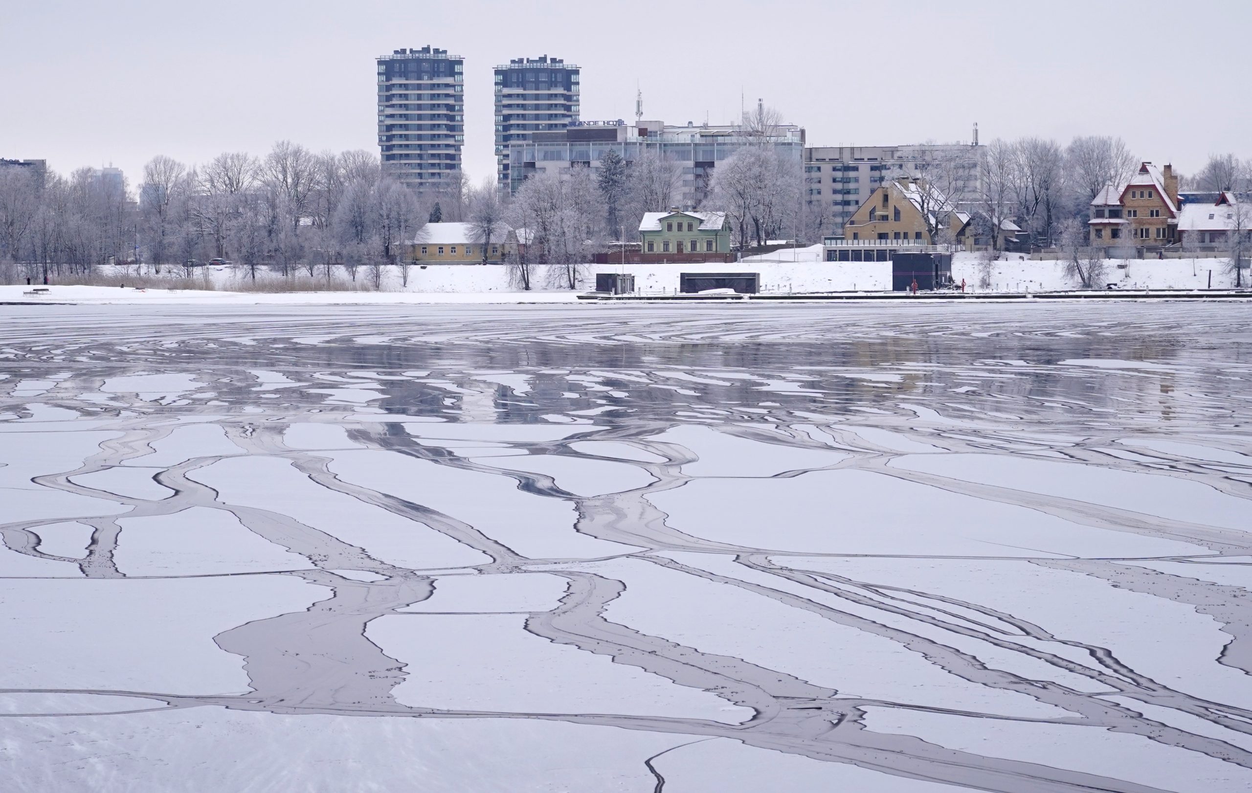 Daugava Rīgā ziemā ar nevienmērīgu ledu un plaisām, fonā pilsētas ēkas un krasts.