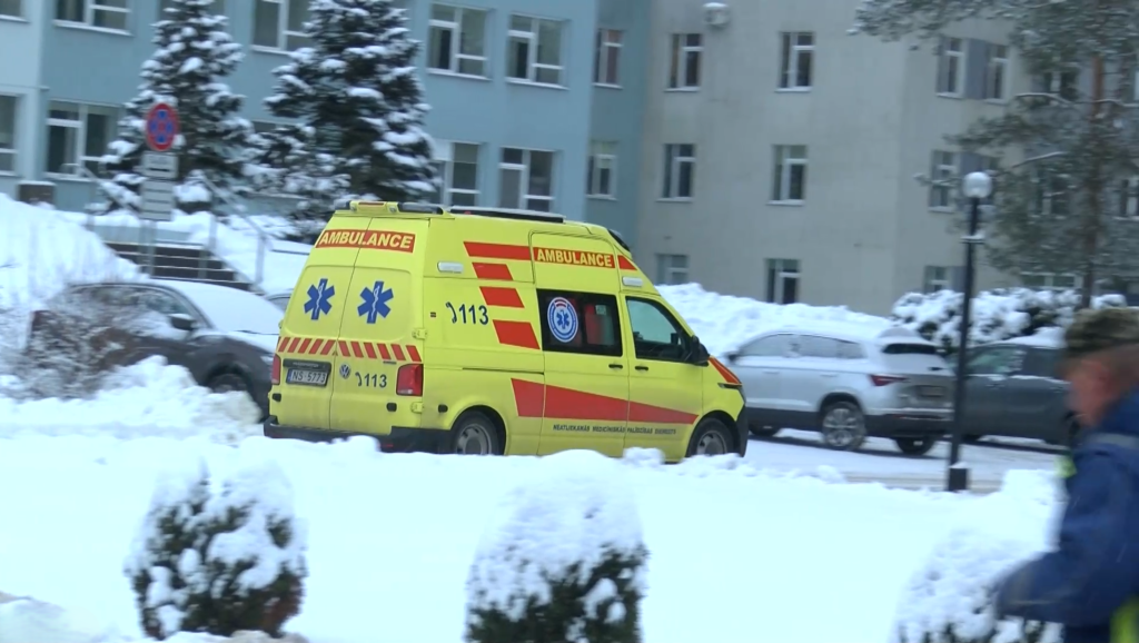 Dzeltena NMPD automašīna ar ieslēgtām bākugunīm brauc pa sniegotu teritoriju pie slimnīcas korpusa.
