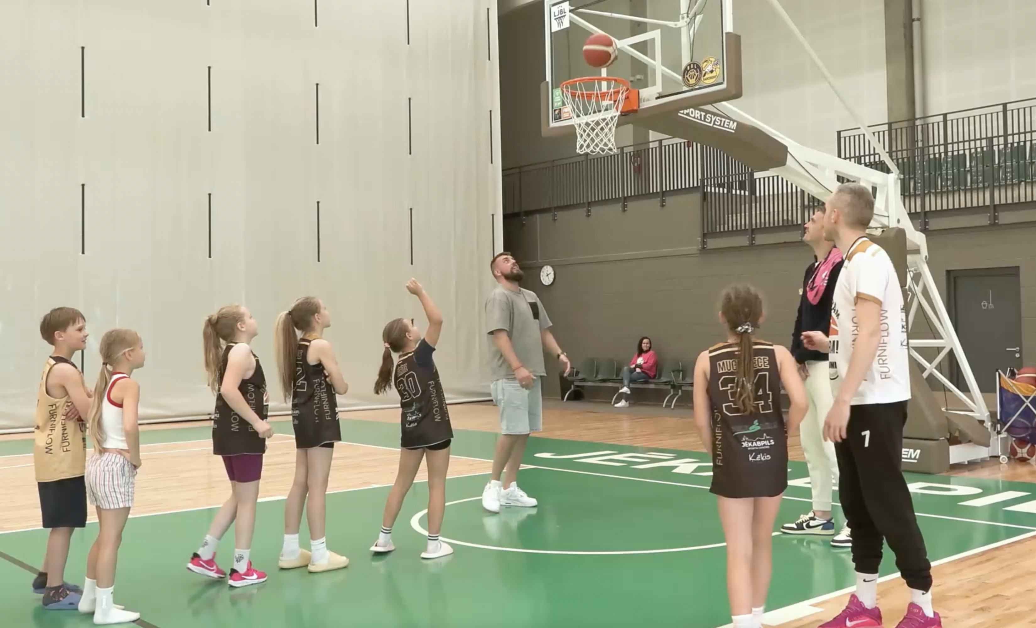 Bērni basketbola treniņā sporta zālē, met bumbu grozā trenera vadībā
