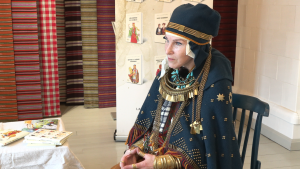 Sieviete latviešu tautastērpā ar bagātīgām rotām sēž izstādes telpā pie grāmatām un austiem audumiem