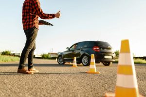 Braukšanas eksāmens autoskolā – instruktors vērtē vadītāja prasmes uz ceļa ar konusiem norobežotā laukumā.