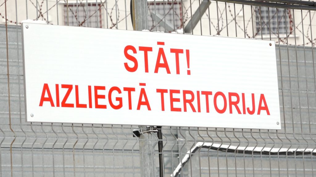 Balta brīdinājuma zīme ar sarkanu uzrakstu "STĀT! AIZLIEGTĀ TERITORIJA" pie dzeloņdrāšu žoga.