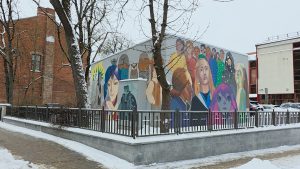 Apgleznots transformatoru skapis Daugavpils ielā ar krāsainu murāli, kurā attēloti dažādi cilvēku portreti un stilizēti tēli.