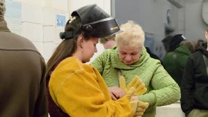 Sieviete metināšanas maskā un aizsargtērpā ar citas personas palīdzību uzvelk dzeltenus aizsargcimdus.