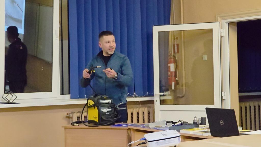 Instruktors demonstrē dzeltenu metināšanas aparātu un tā degli karjeras dienas apmeklētājiem ZRKAC mācību parkā.