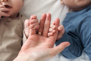 Divi mazuļi tur pieauguša cilvēka pirkstiem, guļot uz gultas mājas apstākļos.