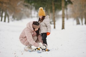 Sieviete rozā ziemas jakā pietupusies palīdz mazai meitenei dzeltenā cepurē nostiprināt slēpju stiprinājumus uz sniegotas nogāzes.