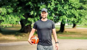Basketbola treneris Oskars Linkovskis ar basketbola bumbu parkā, uzsverot aktīvu un veselīgu dzīvesveidu