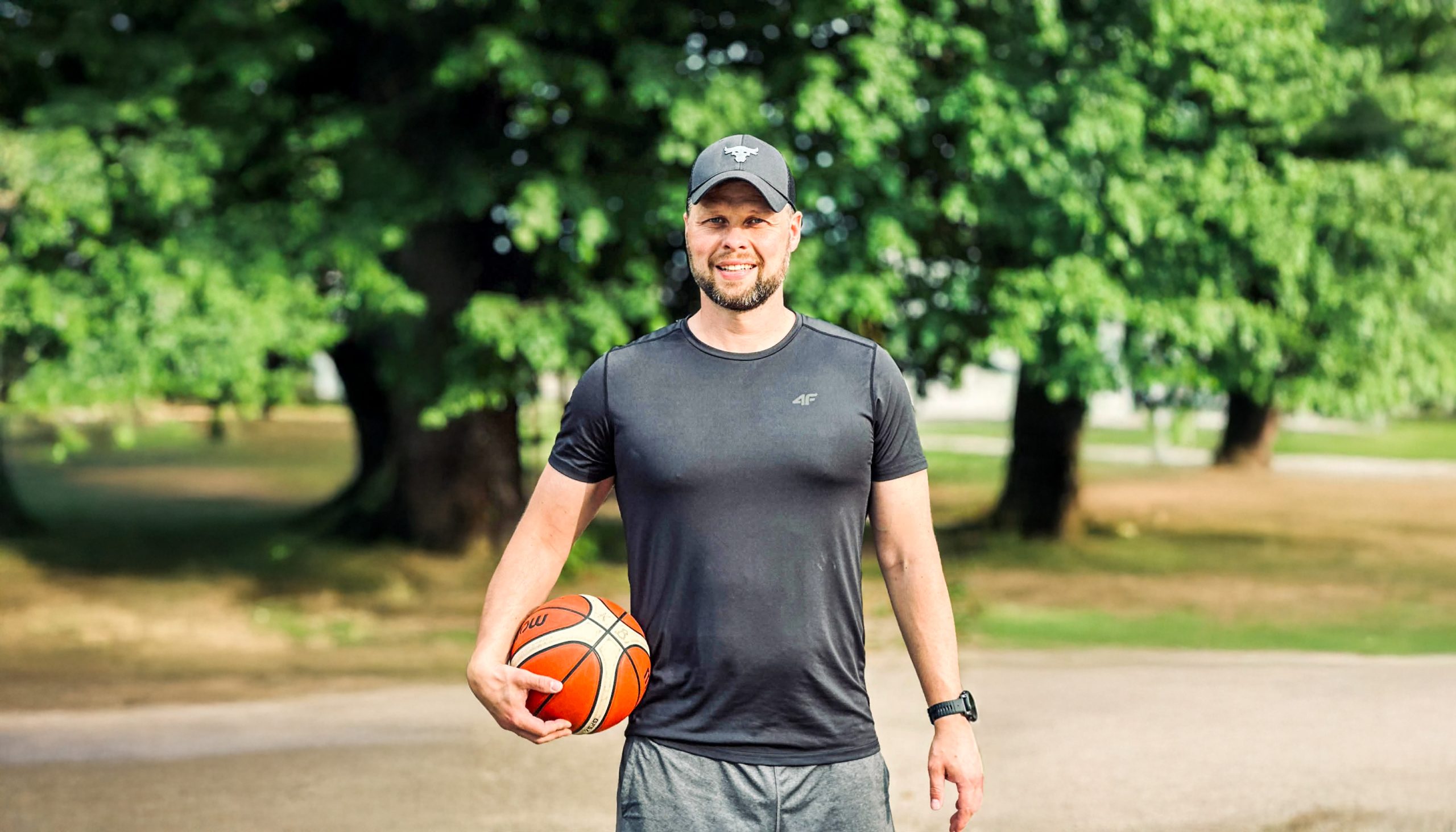 Basketbola treneris Oskars Linkovskis ar basketbola bumbu parkā, uzsverot aktīvu un veselīgu dzīvesveidu