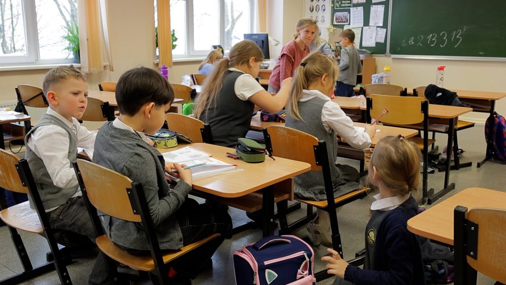 Pirmklasnieki sēž skolas solos un pilda uzdevumus klases telpā pie tāfeles.