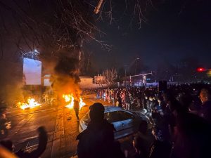 Protestētāju pūlis nakts laikā uz ielas Irānā blakus degošām barikādēm un automašīnai.