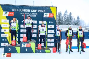 IBU Junioru kausa 2026 apbalvošanas ceremonija Smeceres silā Madonā – biatlonisti uz goda pjedestāla pie sacensību banera.
