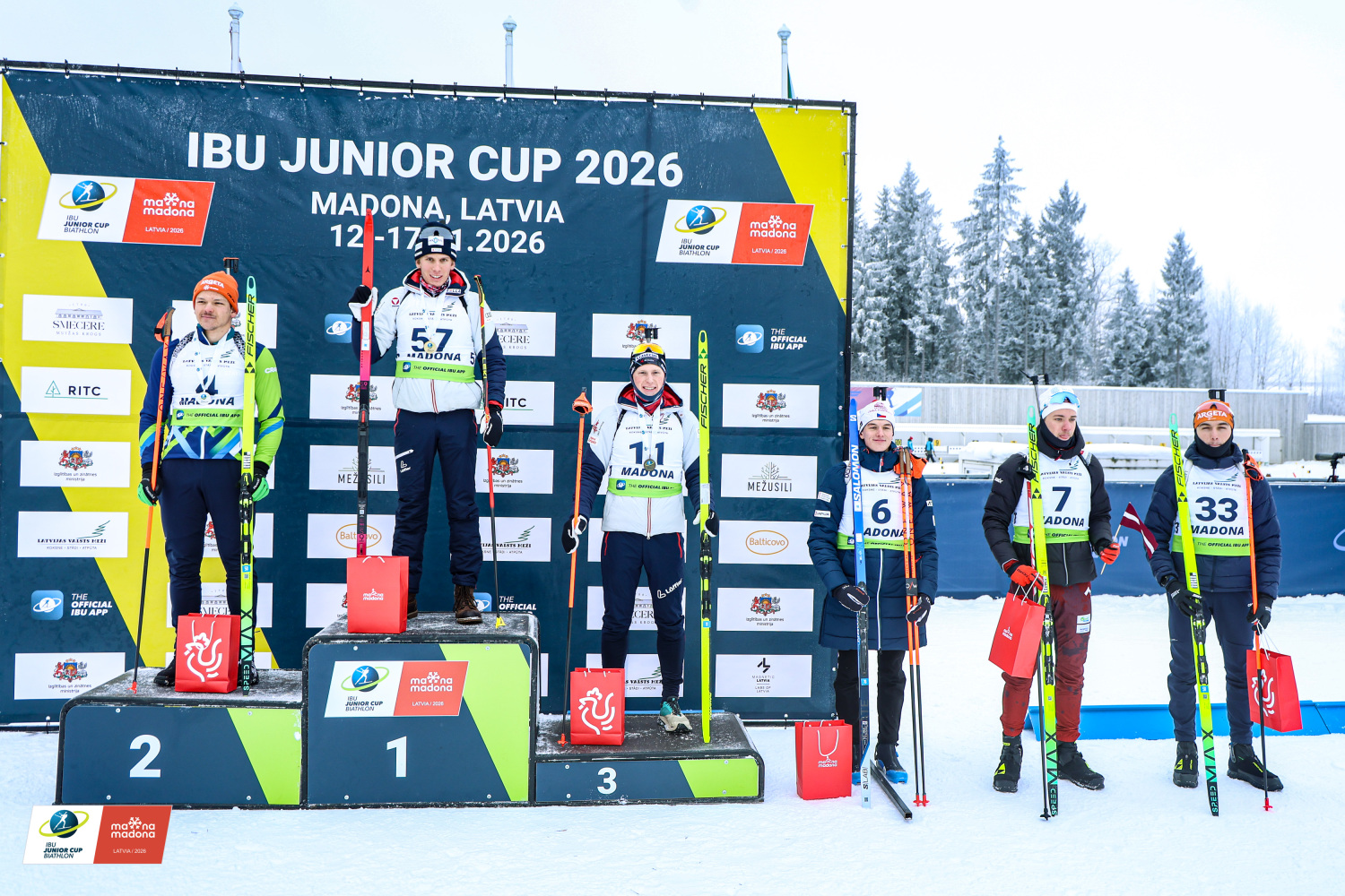 IBU Junioru kausa 2026 apbalvošanas ceremonija Smeceres silā Madonā – biatlonisti uz goda pjedestāla pie sacensību banera.