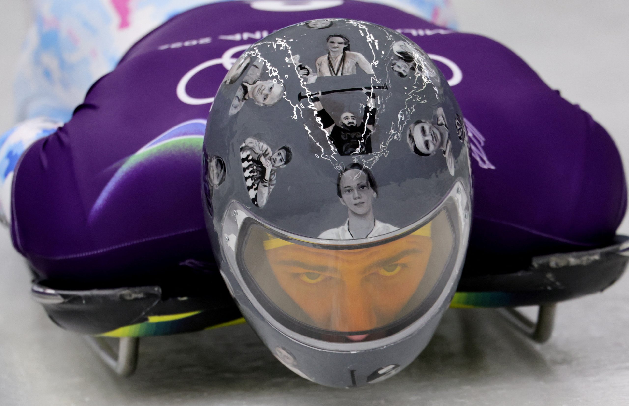 Ukrainas skeletonists Vladislavs Heraskevičs guļ uz skeletona kamanām, skatoties uz priekšu caur ķiveres vizieri, uz kuras redzami nogalināto sportistu attēli.