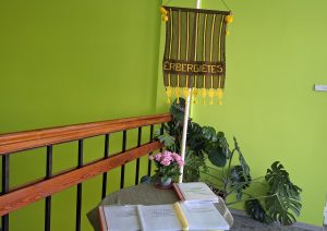 Adīti “Erberģietes” dekori pie zaļas sienas ar telpaugiem un galdu ar grāmatām.