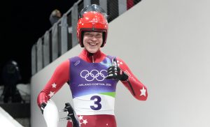 Latvijas kamaniņu sportists ar starta numuru 3 smaida trasē, turot kamanas, gatavojoties braucienam.